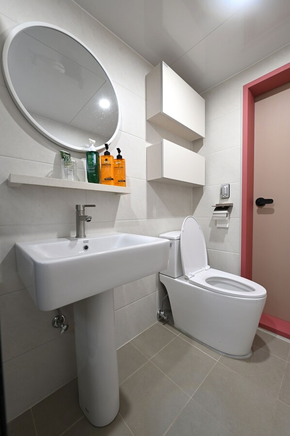 A bathroom with a white sink, toilet, and mirror. There are shelves with toiletries and a red door.