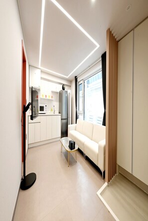Un salon moderne avec un canapé, une table basse et un téléviseur. On aperçoit à l’arrière-plan un coin cuisine avec des placards et des appareils électroménagers.