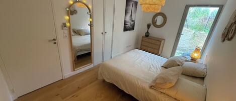 A bedroom with a bed, bedside table, mirror, and a window with a view of greenery.