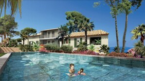 A swimming pool with two people in the water, surrounded by lush greenery and a building with a tiled roof. - Magnifique Appartement Neuf, 3 ch 6 p Centre Ville Avec Jardin Calme et Lumineux (Sainte-Maxime)
