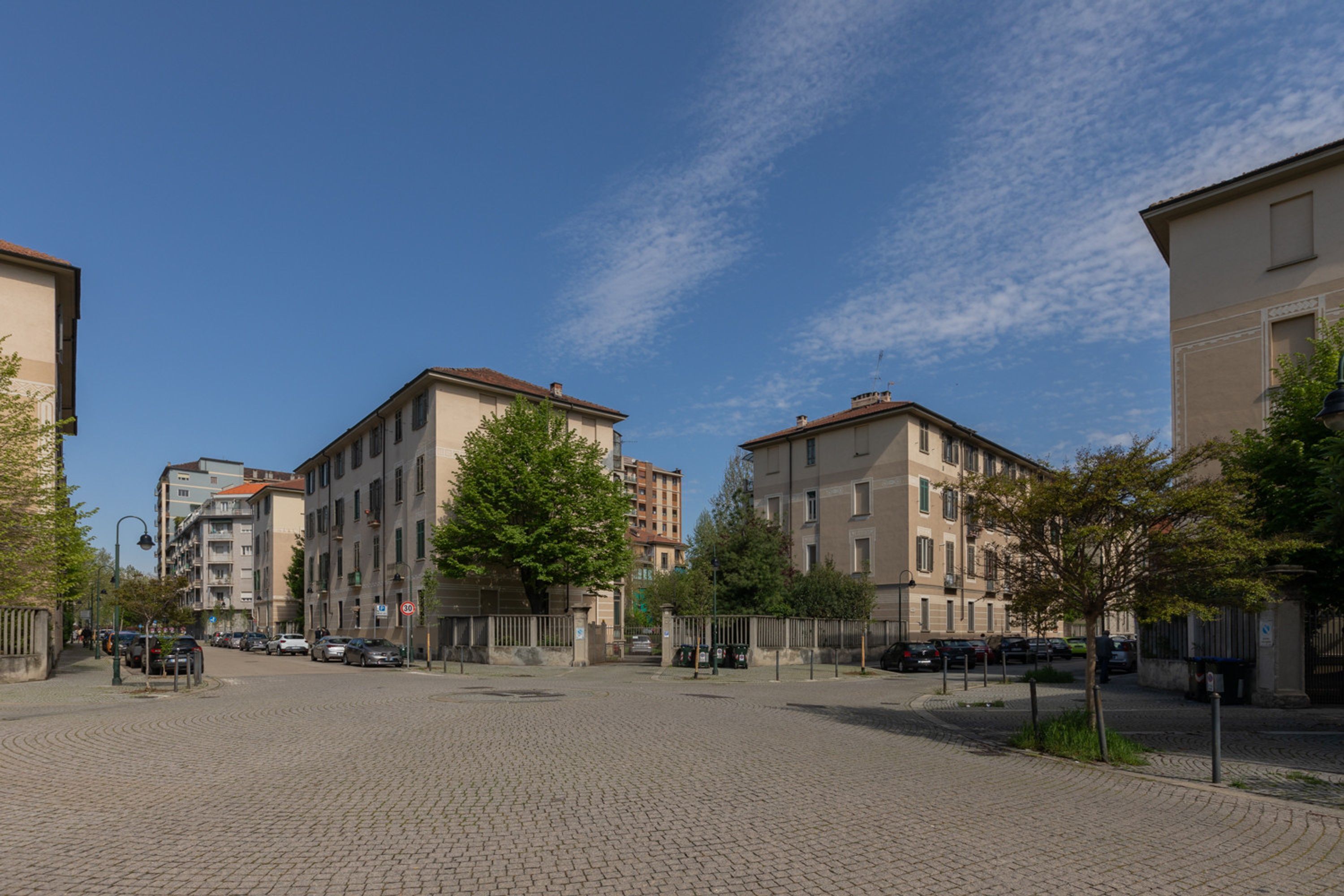 A paved square with buildings on either side, a clear sky, and a few trees.