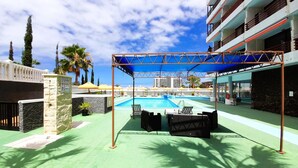 A rooftop pool area with a blue canopy, lounge chairs, and a view of the ocean and buildings in the background. - OLYMPIA STUDIO OCEAN VIEW (Playa de las Américas)