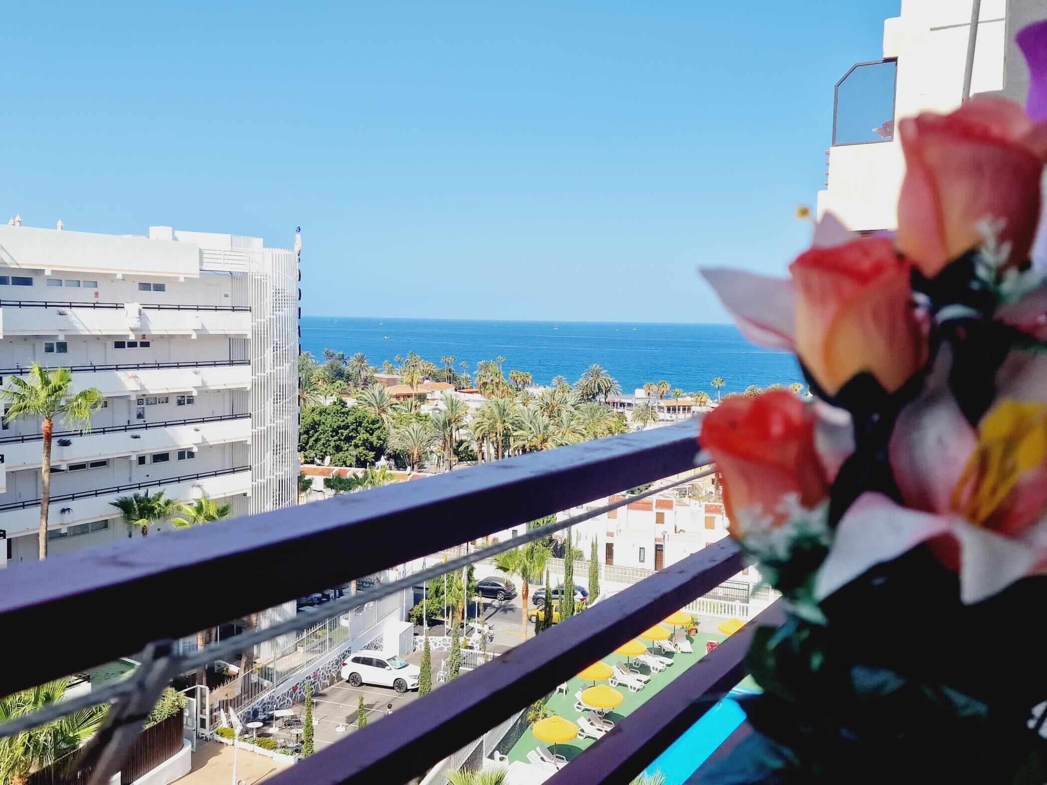 A balcony with a view of the sea, palm trees, and a building with balconies.