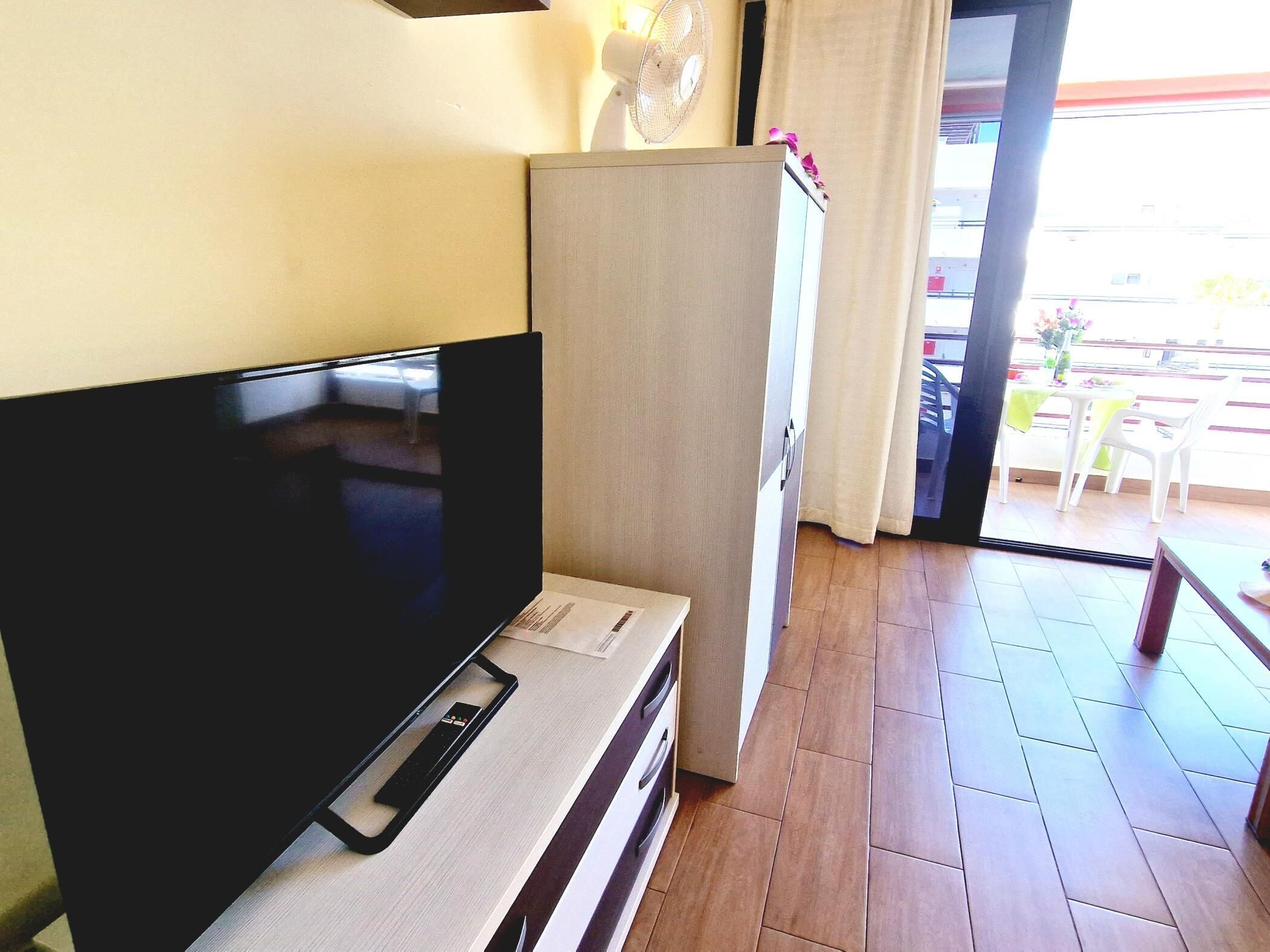 A room with a flat-screen TV, a wooden cabinet, a fan, and a view of the outside balcony with a table and chairs.