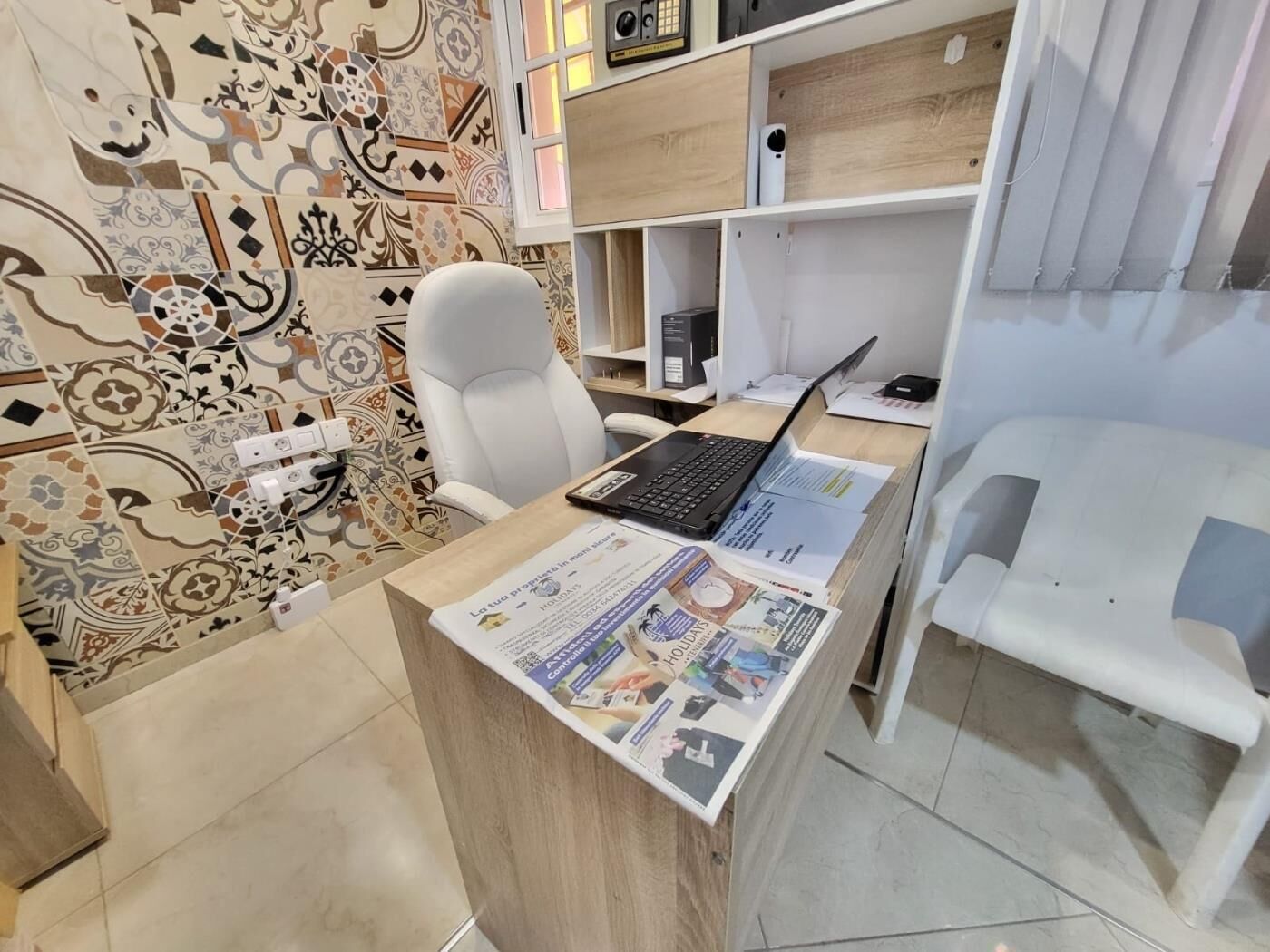 Ein modernes Büro mit einem weißen Schreibtisch, einem Laptop und Unterlagen. Ein weißer Stuhl und ein Regal mit einigen Gegenständen ergänzen das Bild.