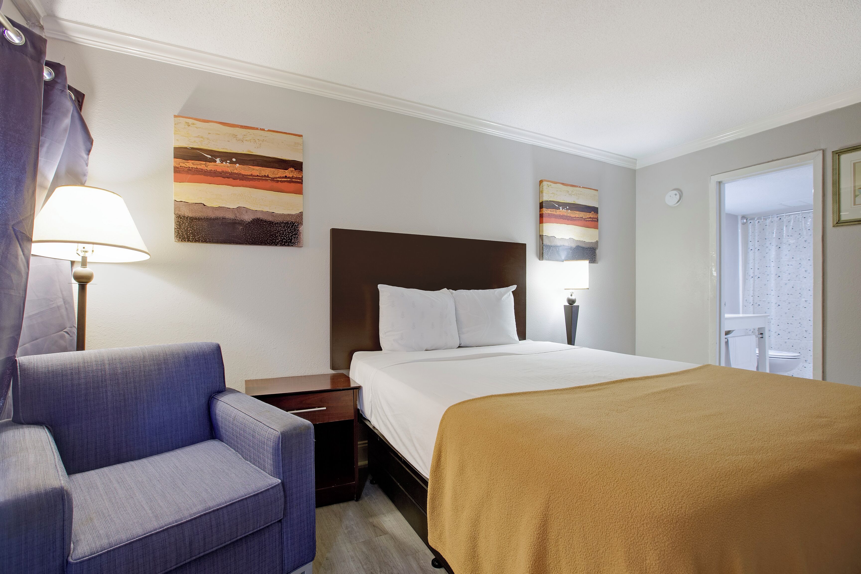 A hotel room with a bed, a chair, a nightstand, a lamp, and two paintings on the wall.