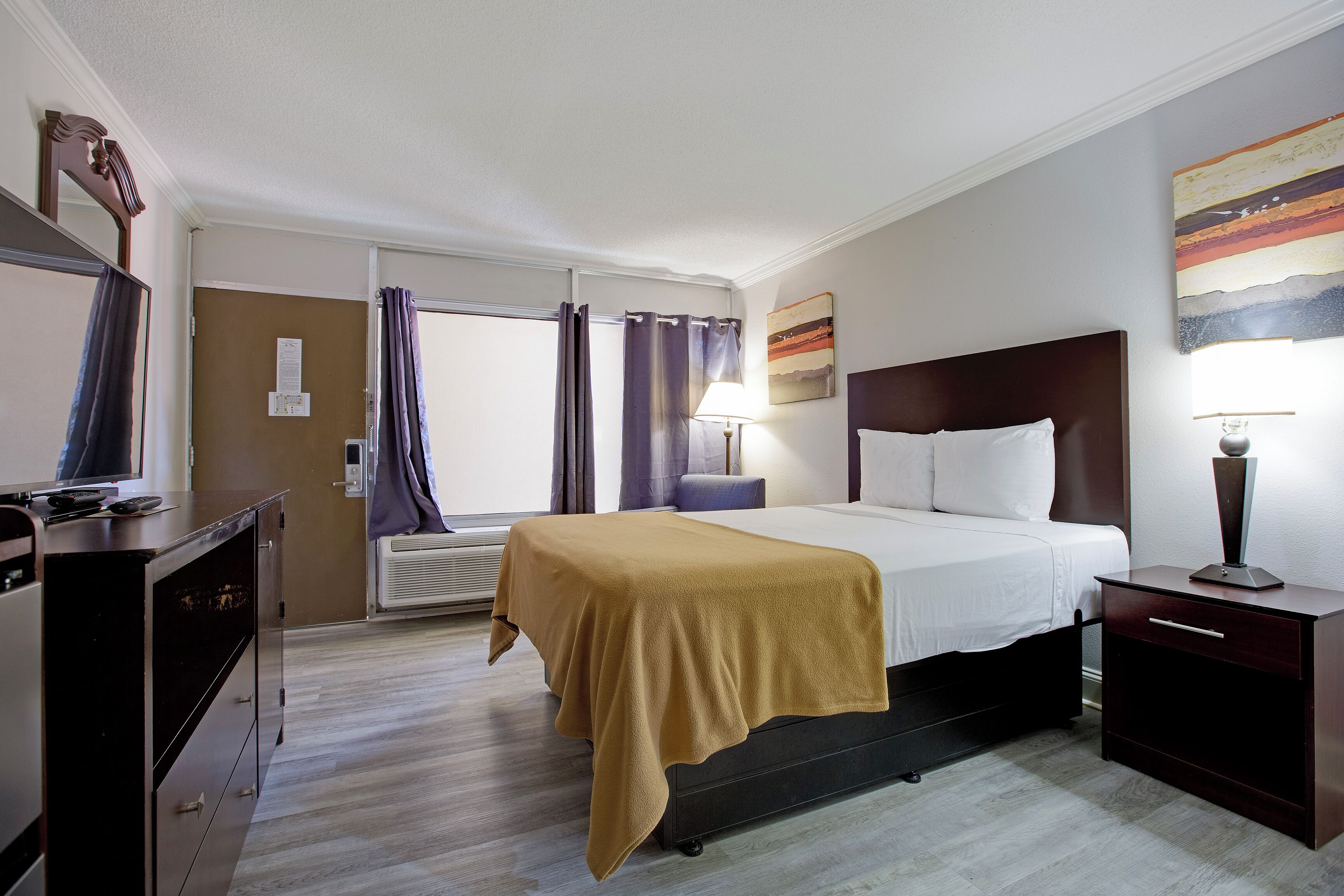 Una habitación de hotel con cama, mesita de noche, lámpara y televisor sobre un tocador.