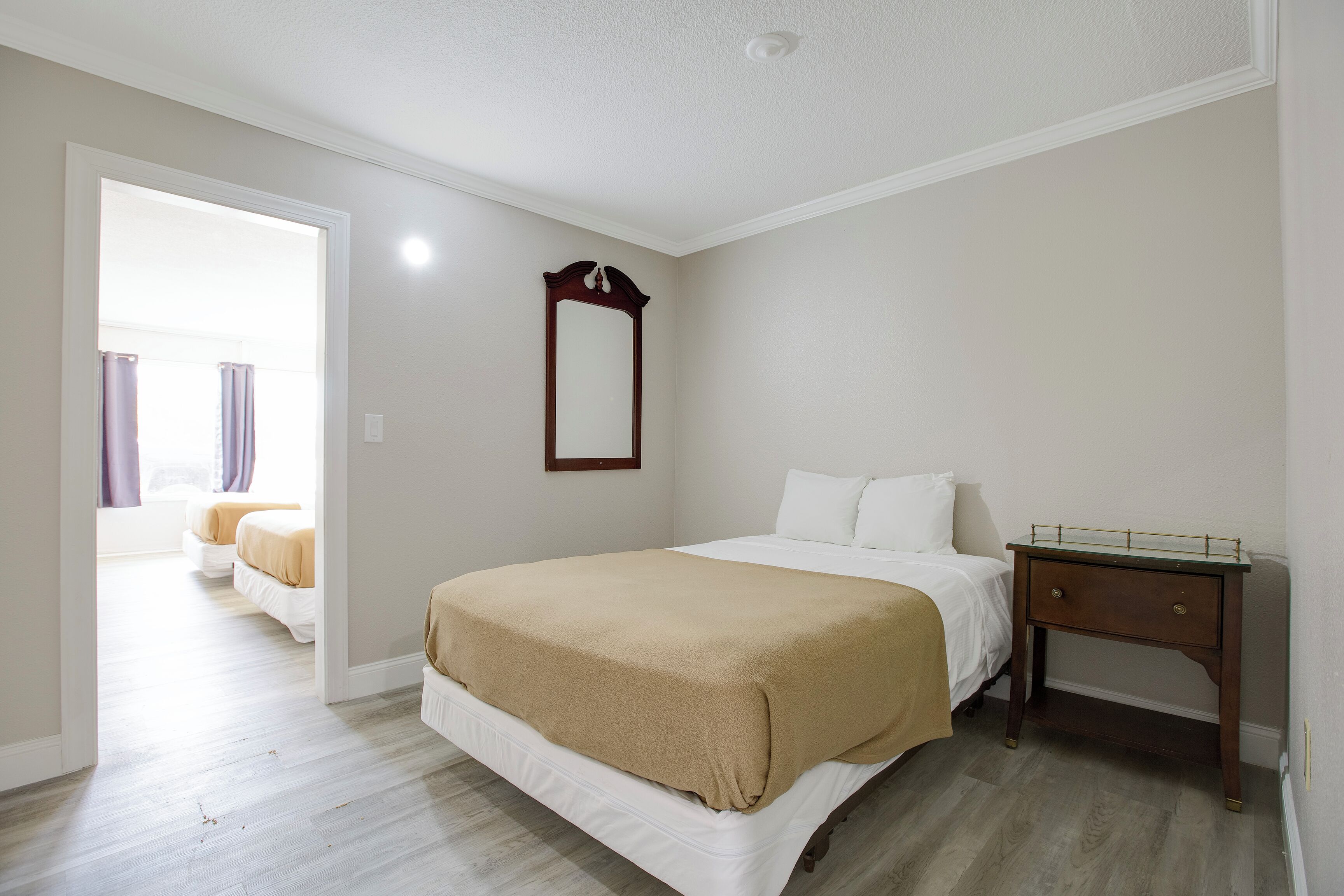 A hotel room with a bed, a bedside table, a mirror, and a view of another room.