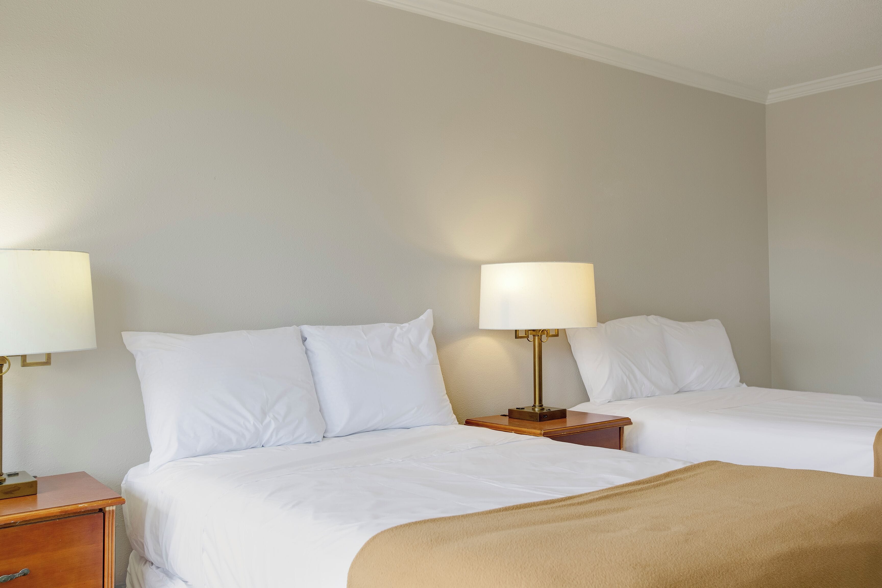 A hotel room with two beds, each with white linens and pillows, a bedside table with a lamp, and a wooden nightstand.