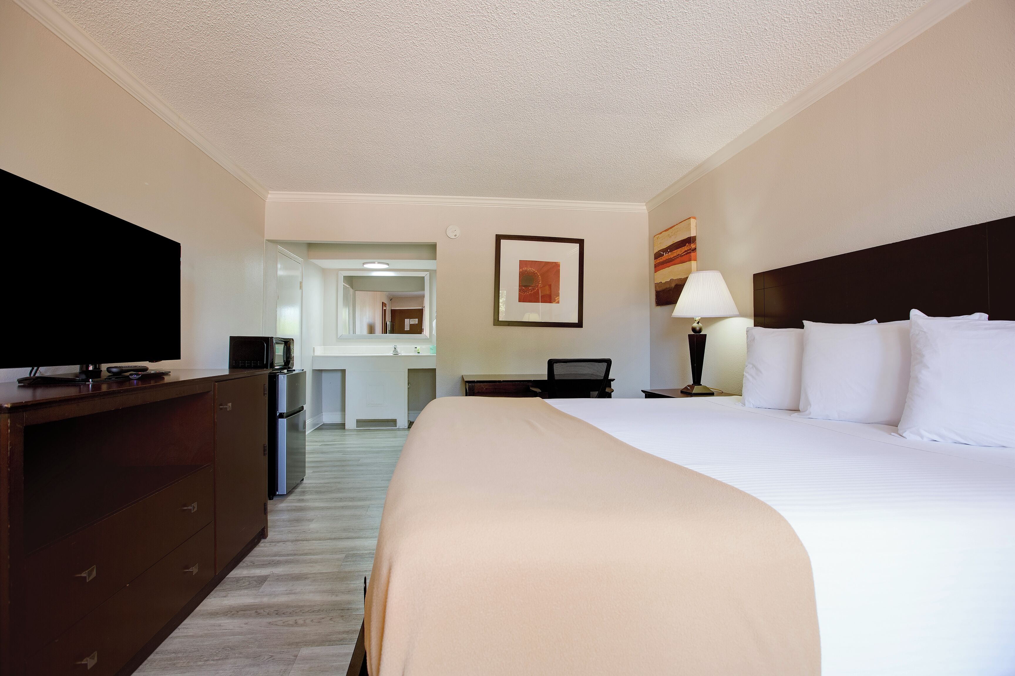 A hotel room with a bed, a television on a dresser, a desk with a lamp, and a painting on the wall.