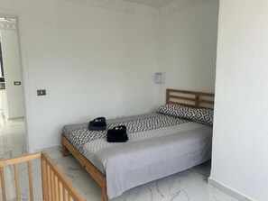 Un lit simple avec une couvre-lit à motifs et des oreillers, une tête de lit en bois, une petite table de chevet et un interrupteur sur le mur.