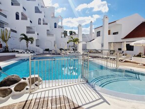 Un espace piscine avec une piscine à l’eau cristalline, des chaises longues et un bâtiment blanc doté de balcons.