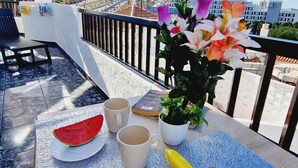 A balcony with a table set for breakfast, including a watermelon, two cups, a banana, and a potted plant with flowers. - Garajonay 3 bedrooms apartment, ocean pool view (Costa de Adeje)