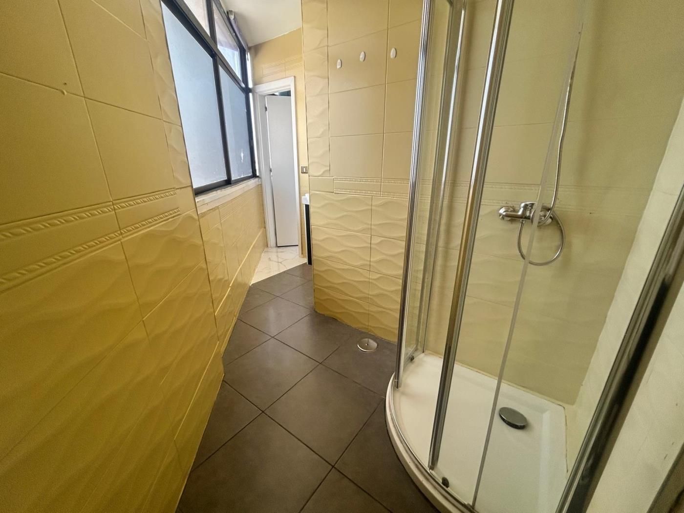 Un bany petit i estret amb plat de dutxa de vidre, parets revestides de rajola i una porta visible que condueix a una altra estança.