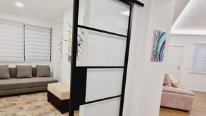 Un salon moderne avec un canapé d’angle, un petit banc et un fauteuil rose. On y trouve un élément décoratif mural et une porte avec un cadre noir.