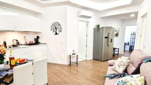 Une cuisine moderne avec des meubles blancs, une table à manger avec des chaises, un réfrigérateur et un coin salon avec un canapé et des coussins décoratifs.