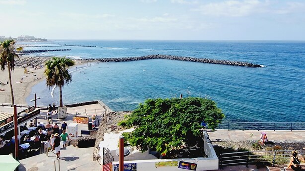 Un restaurant situé en bord de mer, avec une terrasse offrant une vue sur l’océan, une digue rocheuse et un ciel d’un bleu limpide.