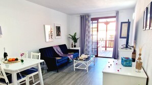 Una sala moderna con un sofá azul, una mesita de centro blanca y una mesa de comedor con sillas. Hay una planta, una botella y un jarrón sobre la mesa.