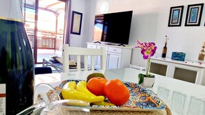 Un comedor con una mesa dispuesta para dos personas, con un frutero que tiene plátanos y naranjas, una copa de vino y un plato decorativo.