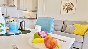 Una cocina moderna con armarios blancos, una silla azul y una mesa de comedor con frutas y una cafetera.