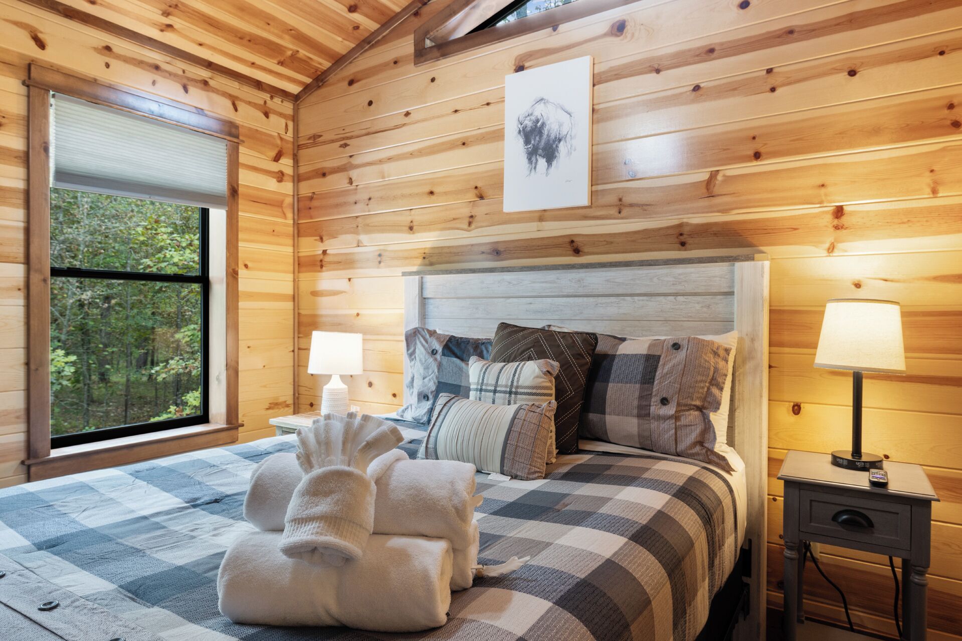 A cozy wooden cabin bedroom with a large bed, two bedside lamps, a framed picture on the wall, and a window overlooking a forest.