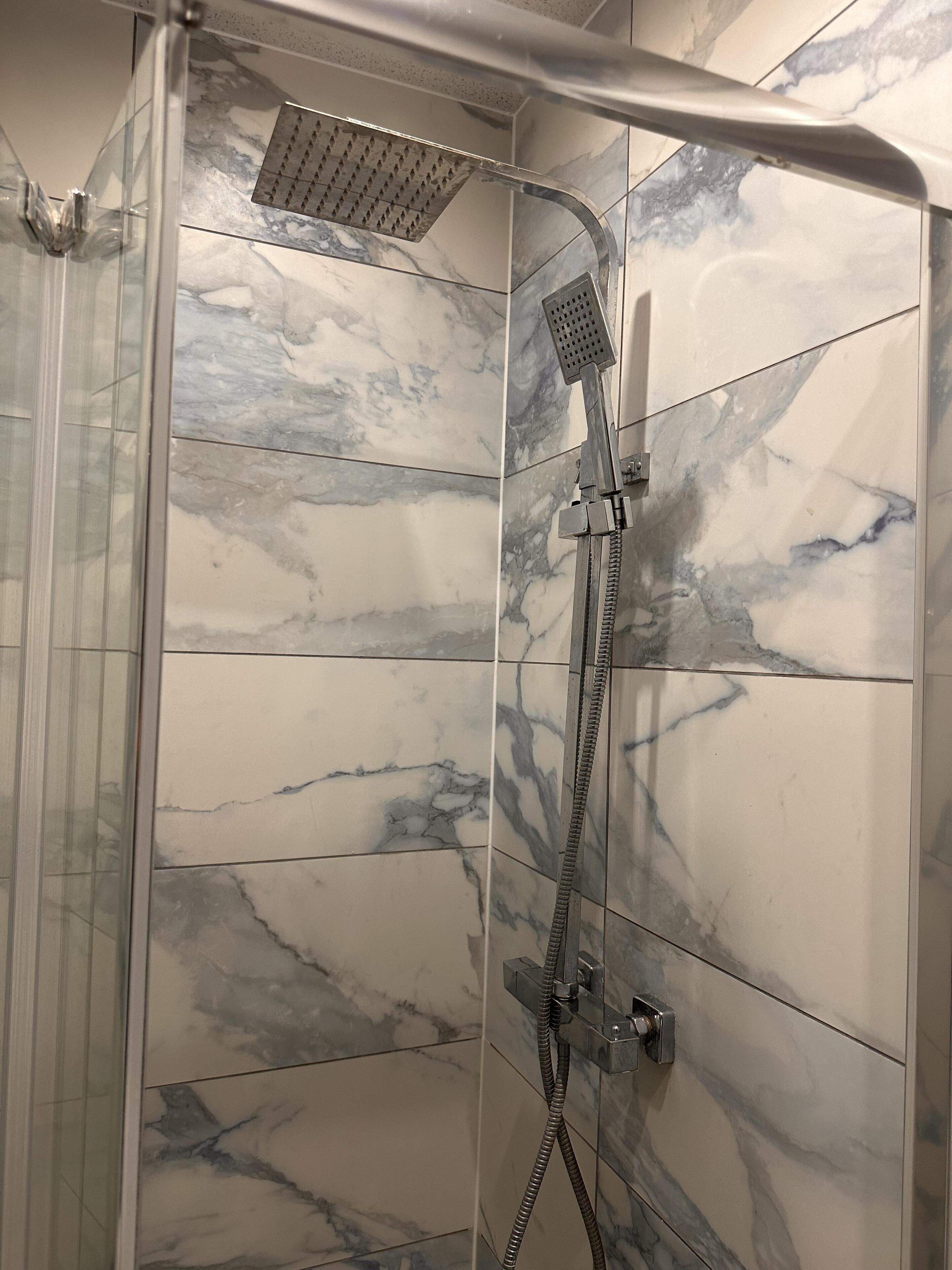 A walk-in shower with a handheld showerhead and a fixed showerhead, both mounted on a wall with marble tiles.