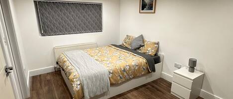 A bedroom with a bed, a bedside table, a nightstand, a window with blinds, and a picture on the wall.