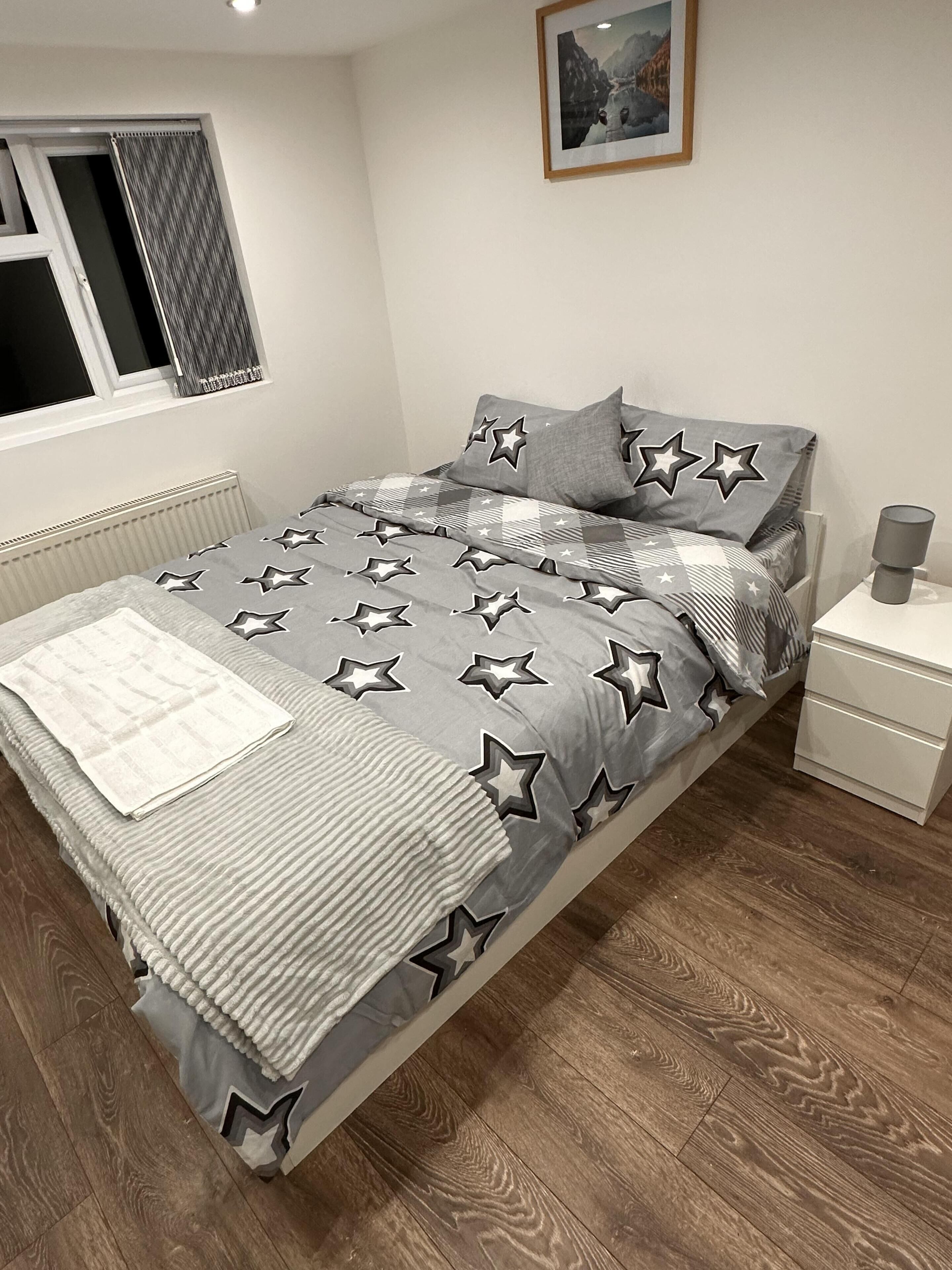 Una cama bien tendida con una colcha estampada de estrellas, una mesita de noche con una lámpara y una ventana con cortinas.