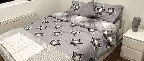 Una cama bien tendida con una colcha estampada de estrellas, una mesita de noche con una lámpara y una ventana con cortinas.