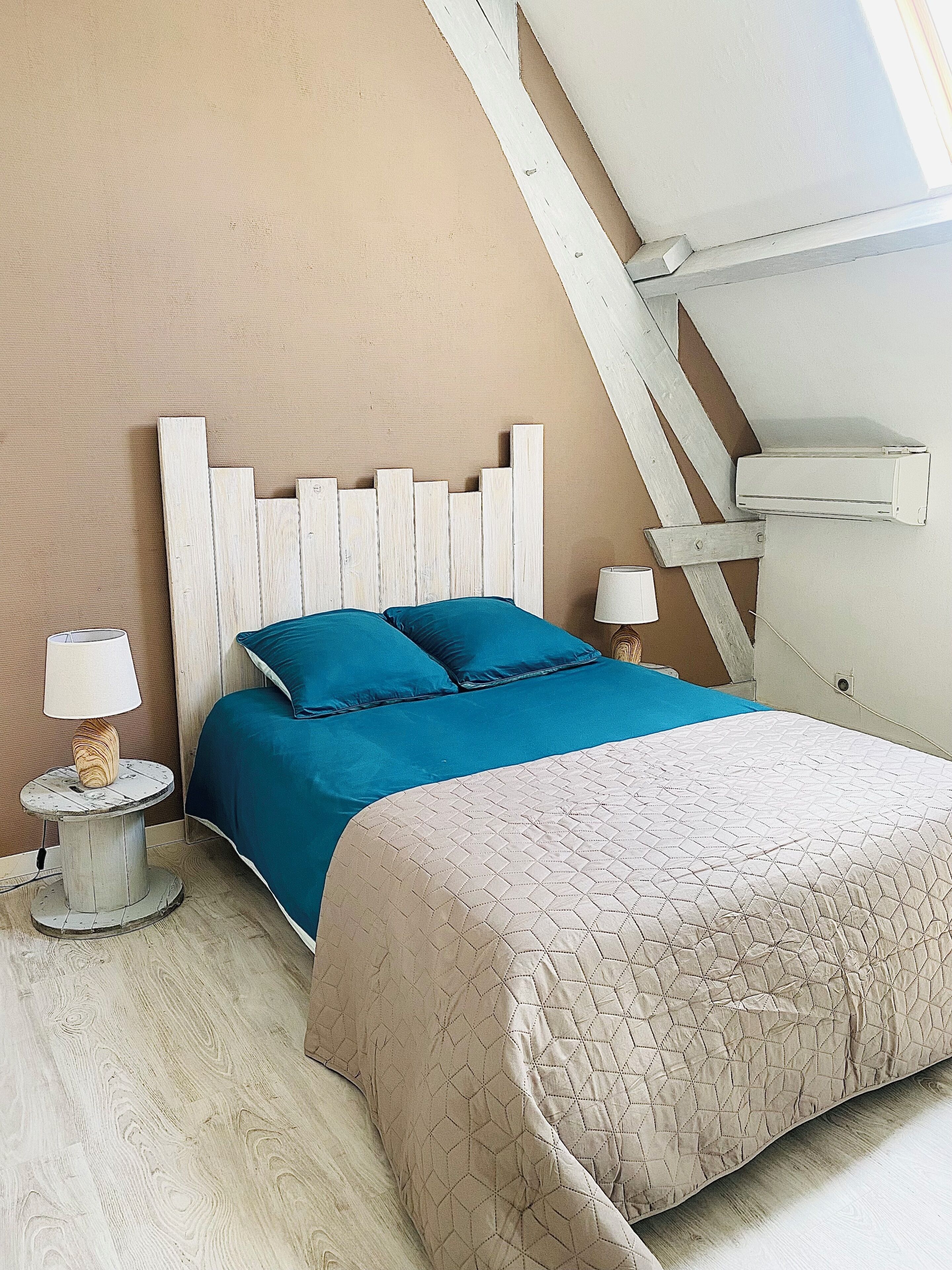 Un dormitorio con cabecera de madera, cama con ropa de cama azul y beige, lámpara de noche y una mesita redonda.