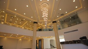 A spacious lobby with a high ceiling, a central chandelier, and a staircase leading to an upper level. - Grand Plaza Hotel (Kirdasah)