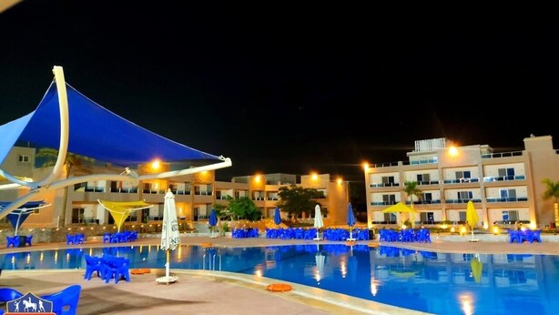 A nighttime view of a hotel pool area with blue lounge chairs and umbrellas, surrounded by buildings with balconies and lights. - Grand Plaza Hotel (Kirdasah)