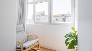 Een kamer met een houten stoel, een potplant en een raam met uitzicht op gebouwen.