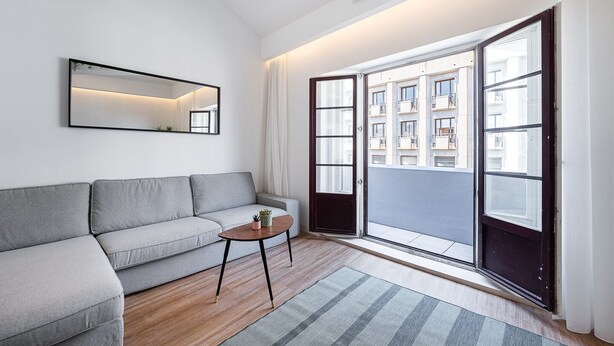 Een moderne woonkamer met een grijze bank, een klein houten bijzettafeltje, een spiegel en een grote schuifpui naar een buitenbalkon.