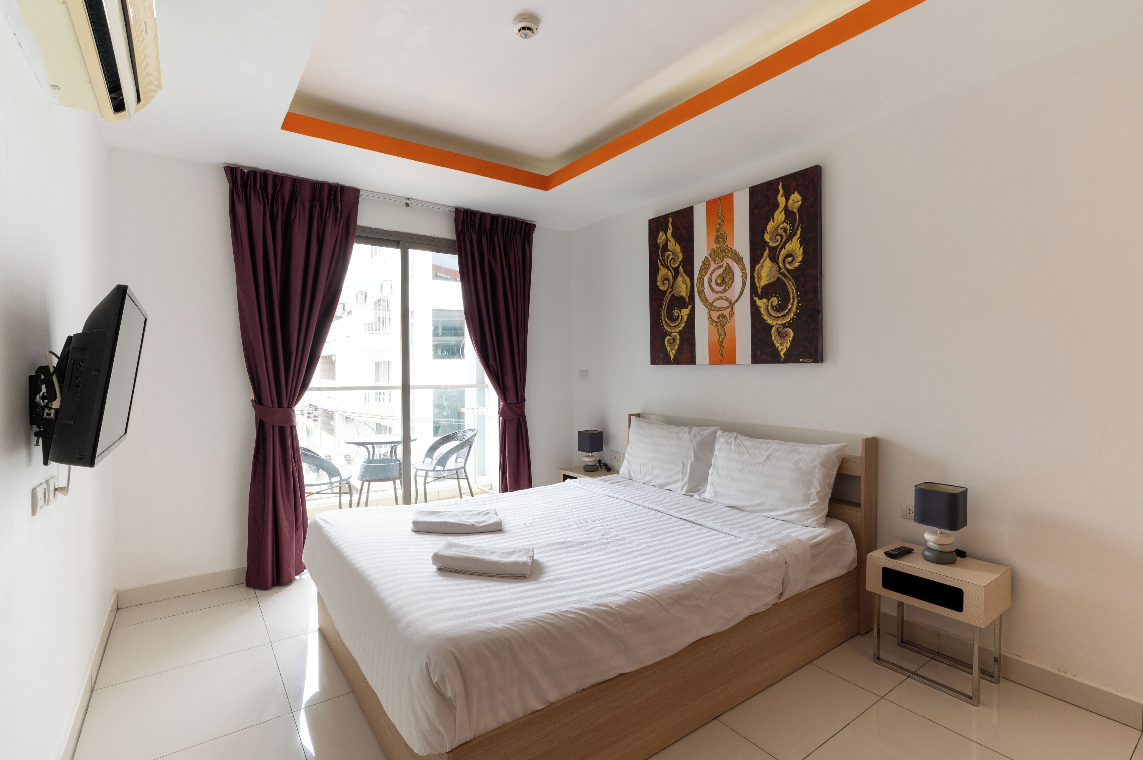 Habitación de hotel con cama, televisor montado en la pared, mesita de noche con lámpara y ventana con cortinas.