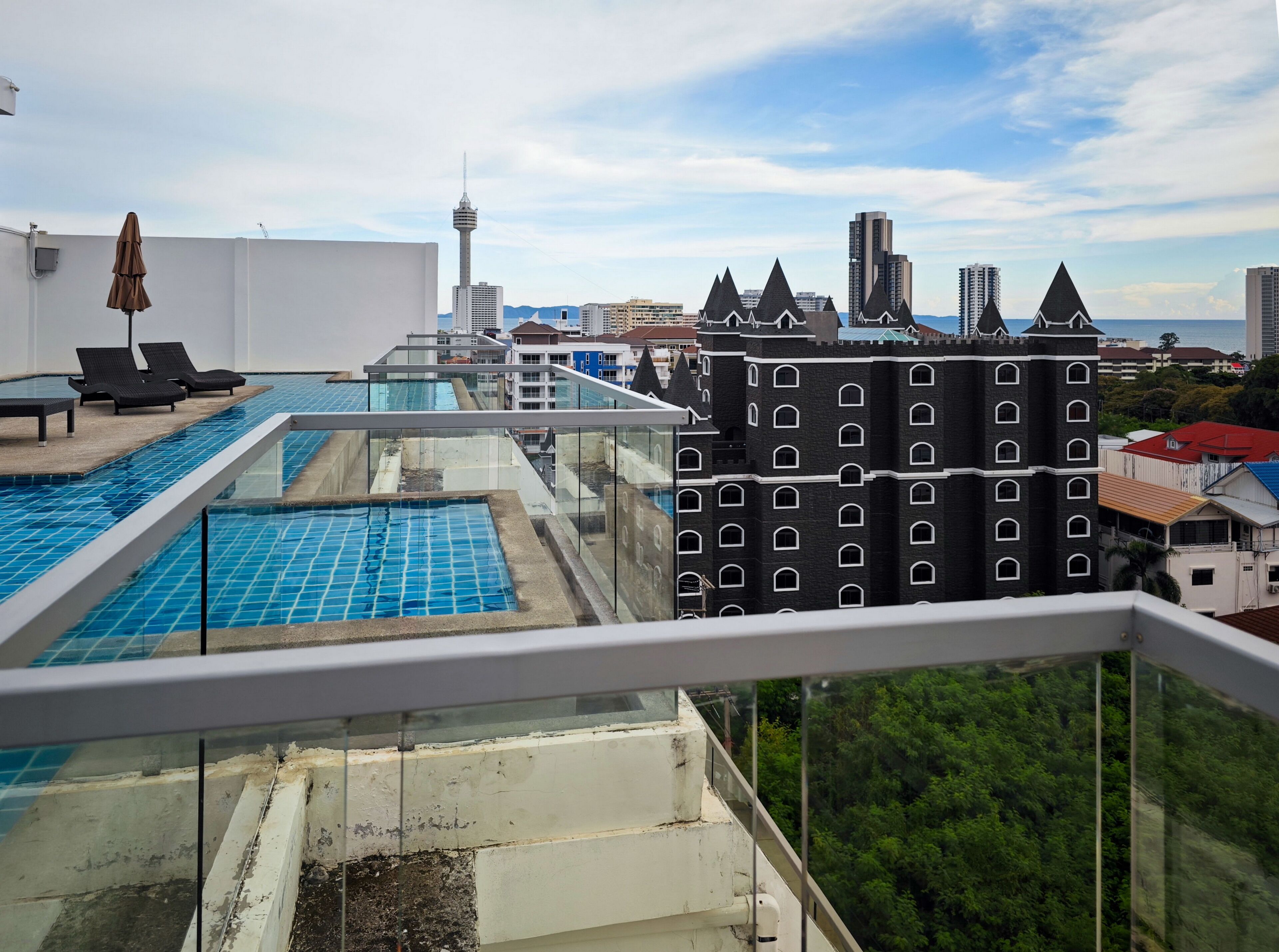 Piscina en la azotea con sillas de descanso y barandilla de vidrio, con vistas a un paisaje urbano de edificios modernos y una torre.