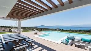 Un espace aménagé au bord de la piscine, avec vue sur la mer, comprenant un fauteuil suspendu et une table avec des coussins.