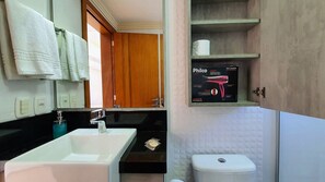 A bathroom with a white sink, a mirror, and a toilet. There is a shelf with a box of toiletries and a roll of toilet paper. - Residencial Dona Virginia (Canela)