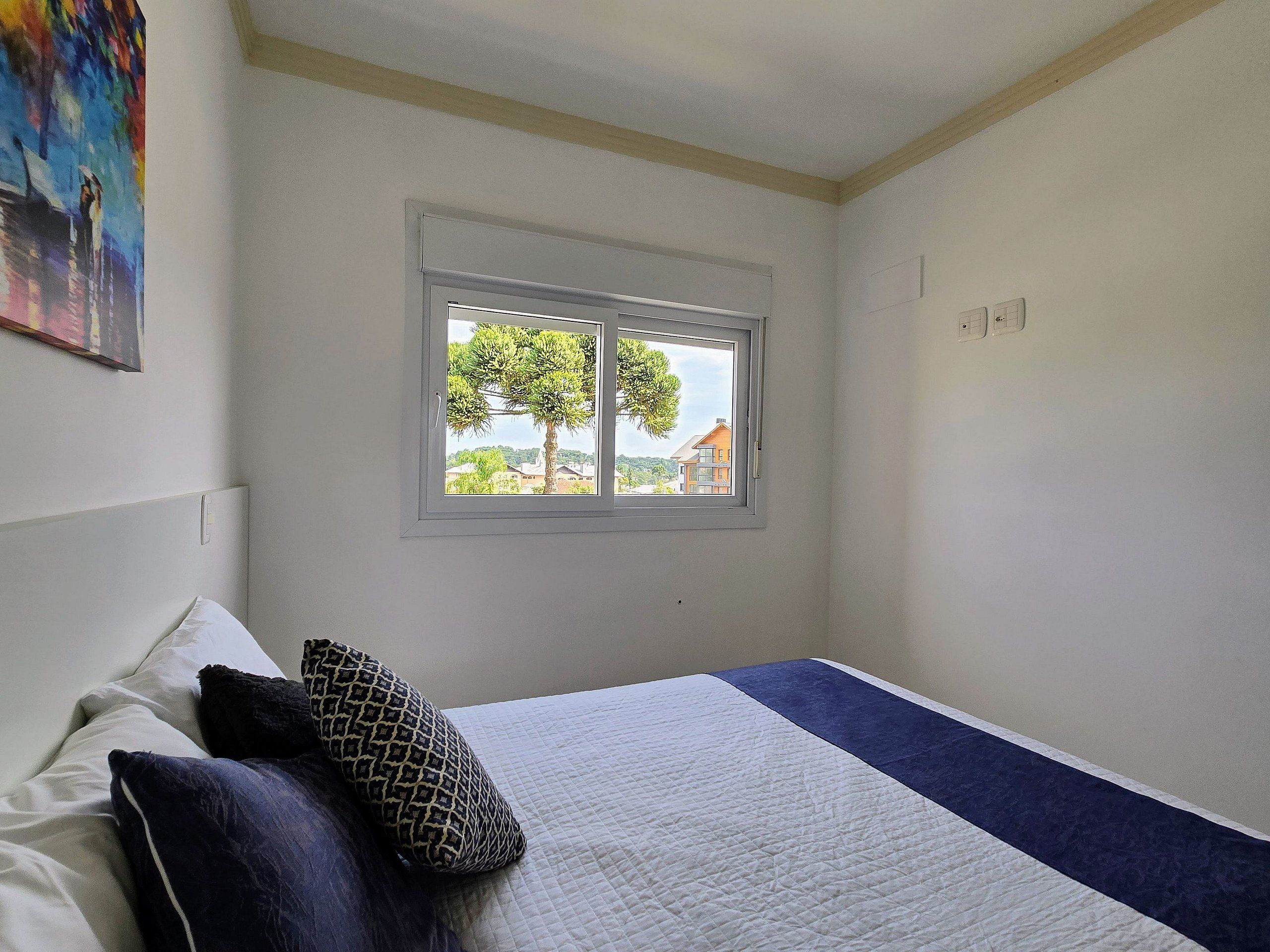 Ein Schlafzimmer mit Bett, Kissen, einem Fenster mit Blick auf Bäume und Häuser sowie einem Bild an der Wand.
