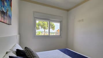 Ein Schlafzimmer mit Bett, Kissen, einem Fenster mit Blick auf Bäume und Häuser sowie einem Bild an der Wand.