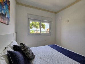 A bedroom with a bed, pillows, a window with a view of trees and houses, and a painting on the wall. - Residencial Dona Virginia (Canela)