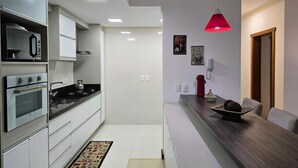 A modern kitchen with white cabinets, a built-in oven, a microwave, and a stove. There is a wooden dining table with a red pendant light above it. - Residencial Dona Virginia (Canela)