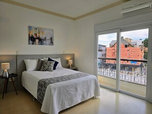 A bedroom with a bed, two bedside tables, a nightstand, a window with a view of buildings, and an air conditioning unit. - Residencial Dona Virginia (Canela)