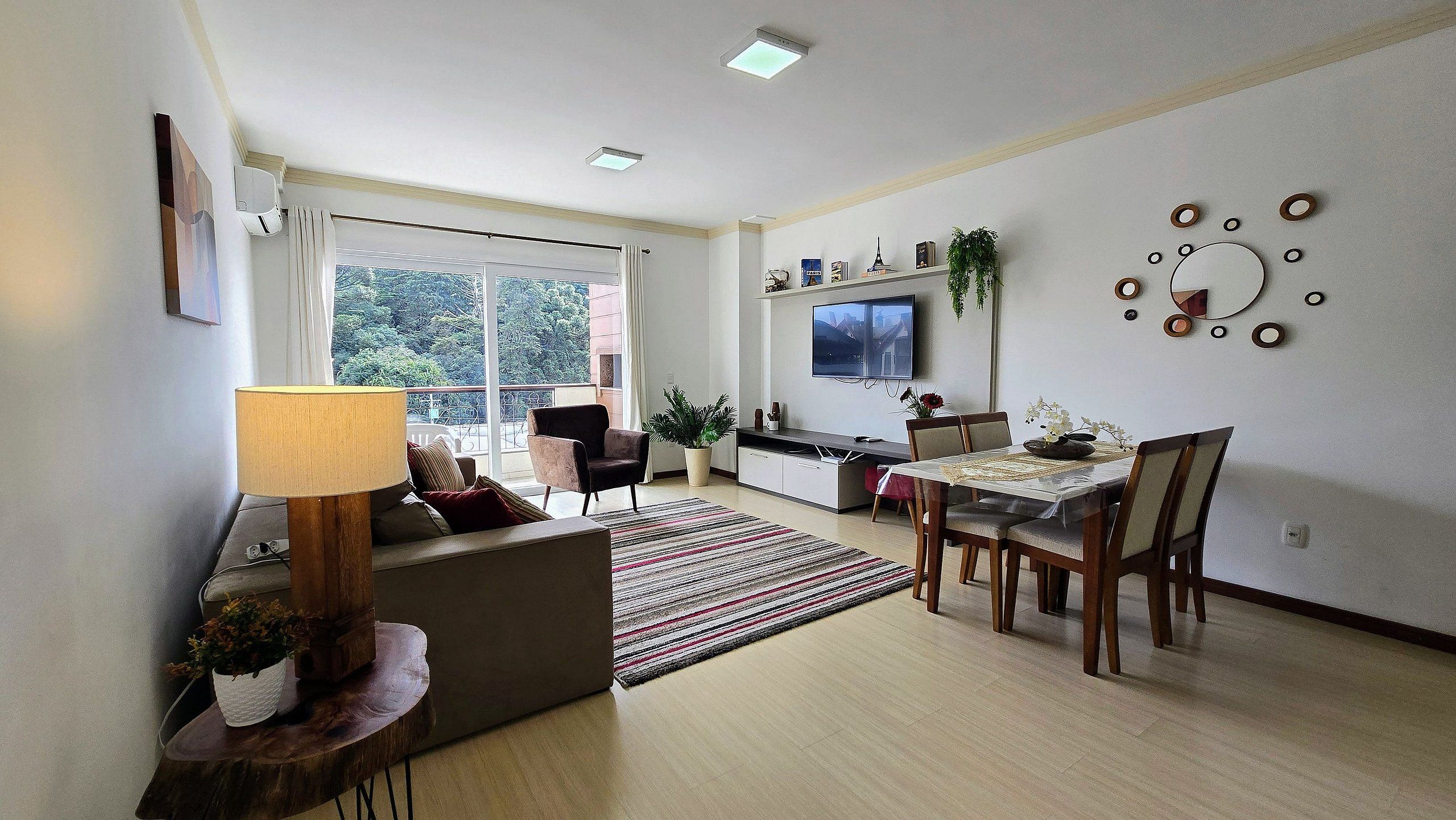 Una sala de estar con un sofá, una mesa de comedor con sillas, un televisor montado en la pared y una ventana con vista a árboles.