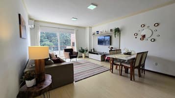 Una sala de estar con un sofá, una mesa de comedor con sillas, un televisor montado en la pared y una ventana con vista a árboles.