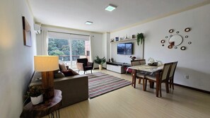 Una sala de estar con un sofá, una mesa de comedor con sillas, un televisor montado en la pared y una ventana con vista a árboles.