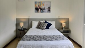 A neatly made bed with a patterned bedspread, two bedside lamps, and a cityscape painting on the wall. - Residencial Dona Virginia (Canela)