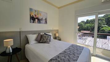 A hotel room with a bed, a painting on the wall, a balcony with a view, and a lamp on a side table.