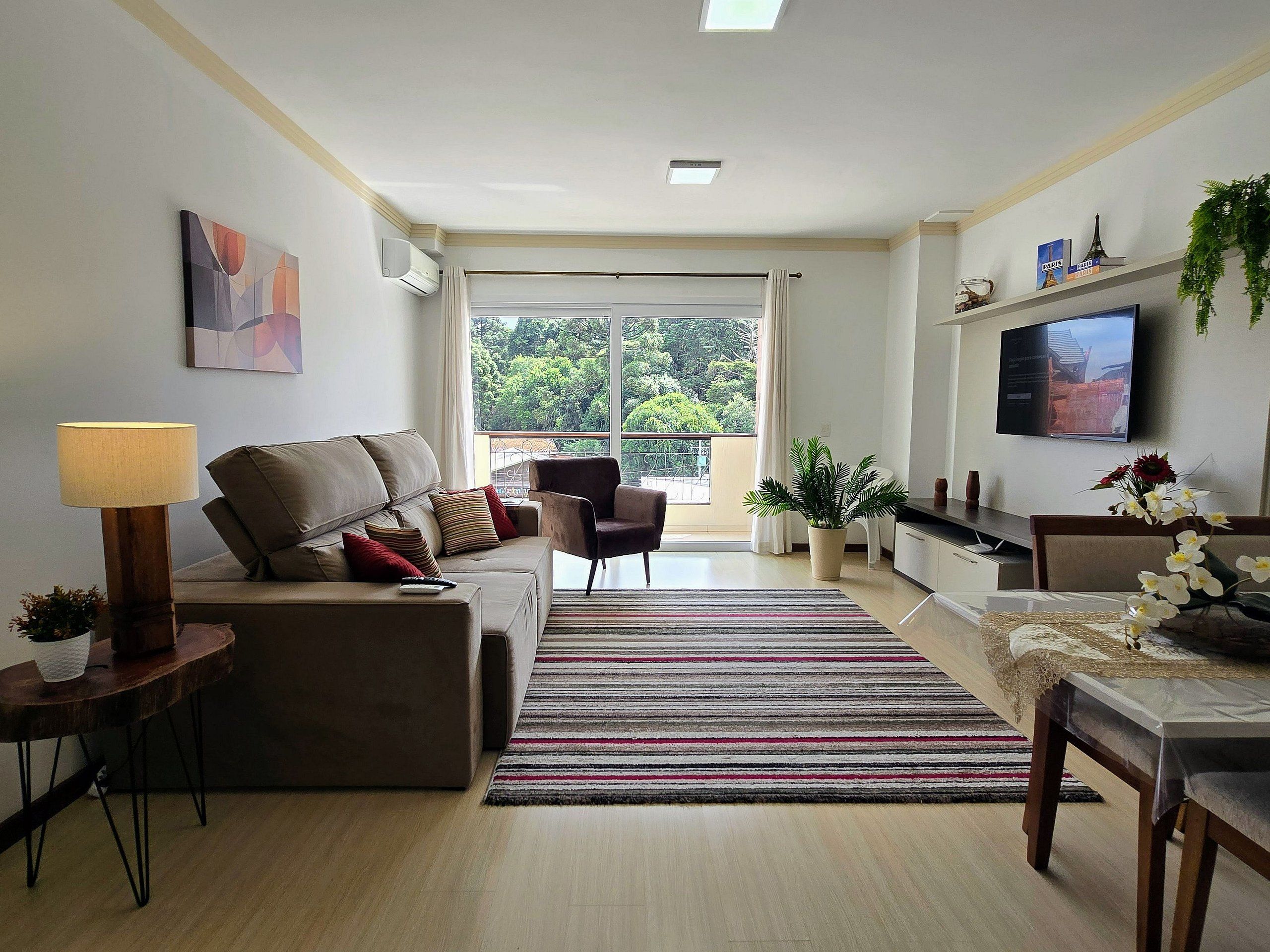 A modern living room with a sofa, armchair, and a dining table with chairs. There is a TV mounted on the wall and a window with a view of greenery.
