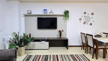 Een woonkamer met een flatscreen televisie, een salontafel, een eettafel met stoelen, een gestreept vloerkleed en een plant.